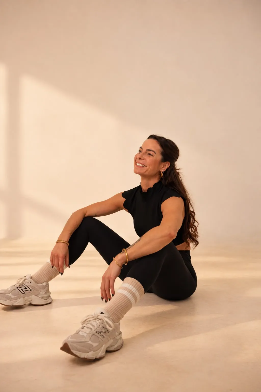 Lucile assise au sol, souriante, en tenue de sport noire, dans un studio baigné de lumière chaude.
